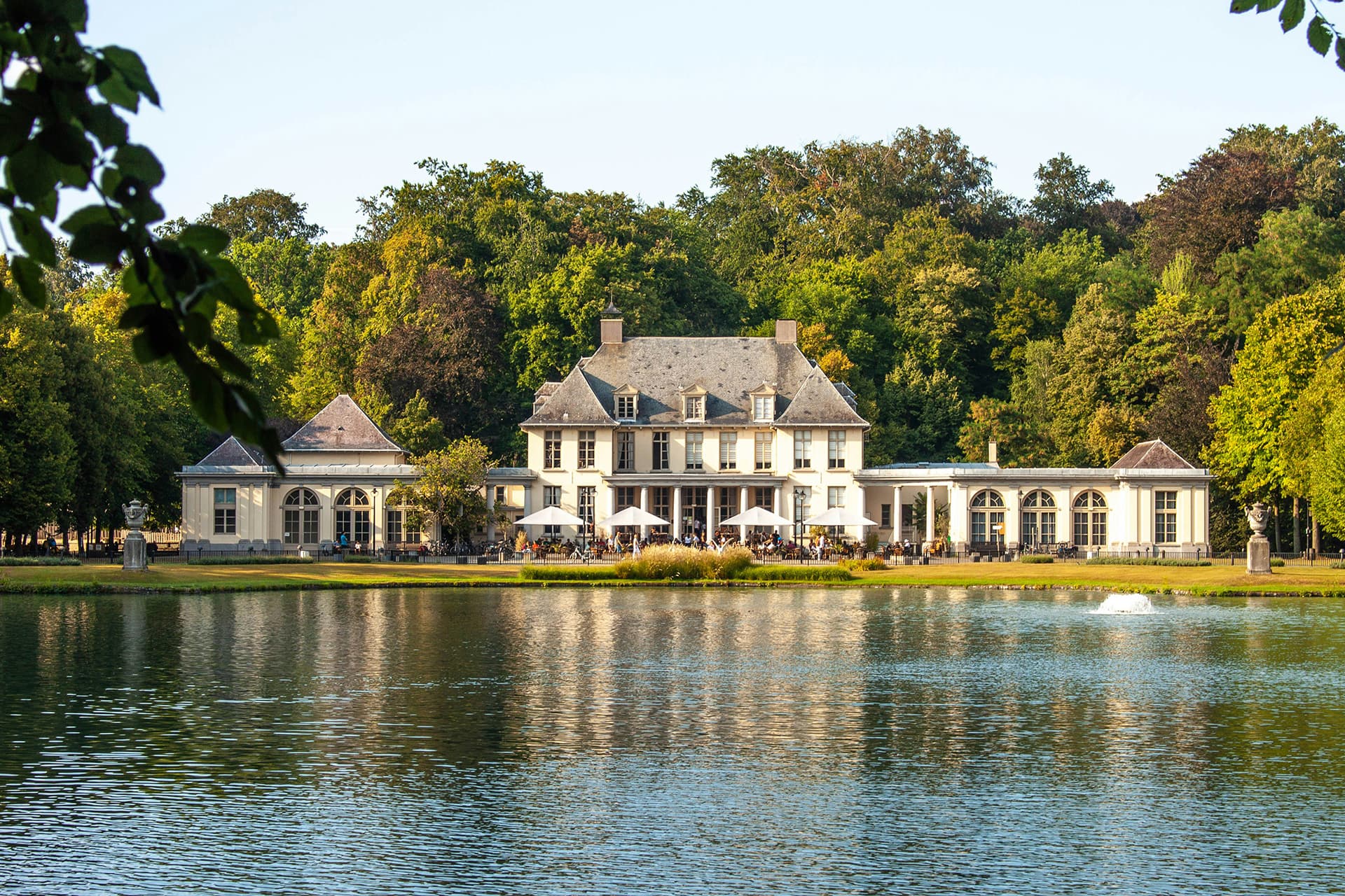 Old, big white building in middle of a park with body of water in front