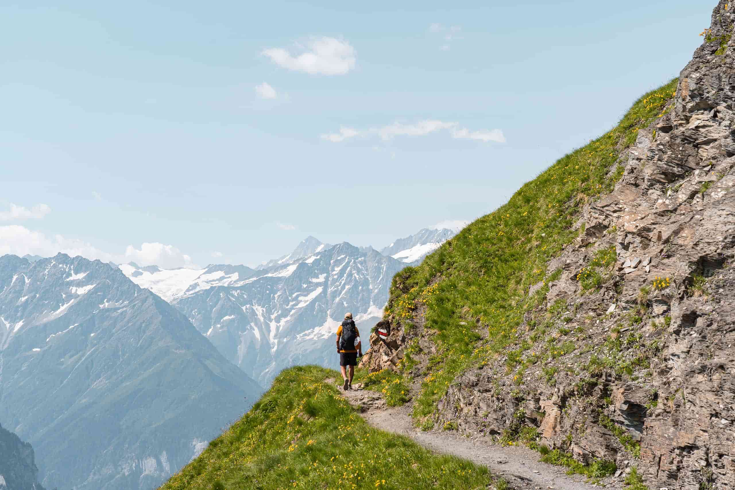 man hiking on small mountain trail with snow covered mountains in the background